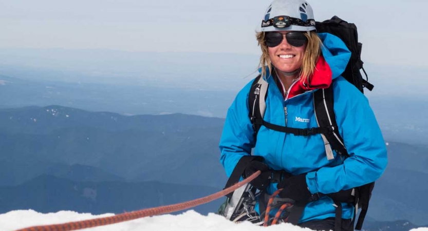 A person wearing mountaineering gear is secured by a rope as they stand on a snowy cliff and smile at the camera. There is a vast mountainous landscape in the background.